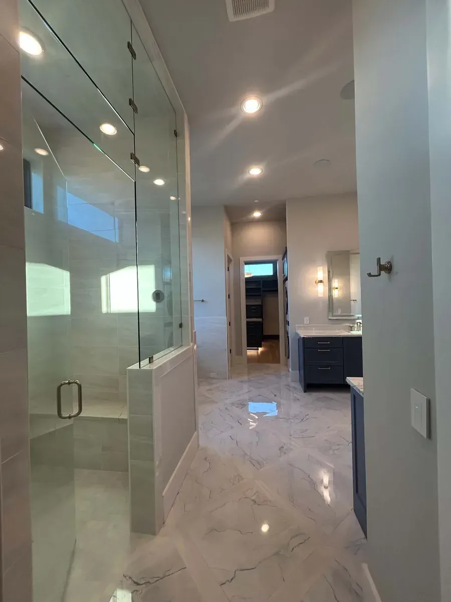 Primary suite walk in shower with frameless glass, marble look floors and a navy vanity beyond